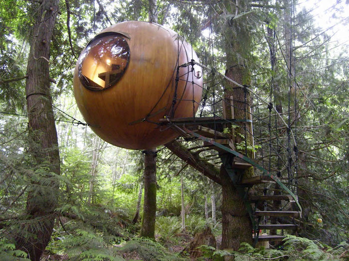 sphere made of wood, with a circular window, suspended from several fir trees, and accessible through a set of stairs