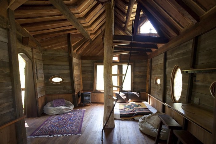 bean bag chairs, ornamental carpet and a bed, inside a boho-style tree house, with several windows, and ceiling with wooden beams