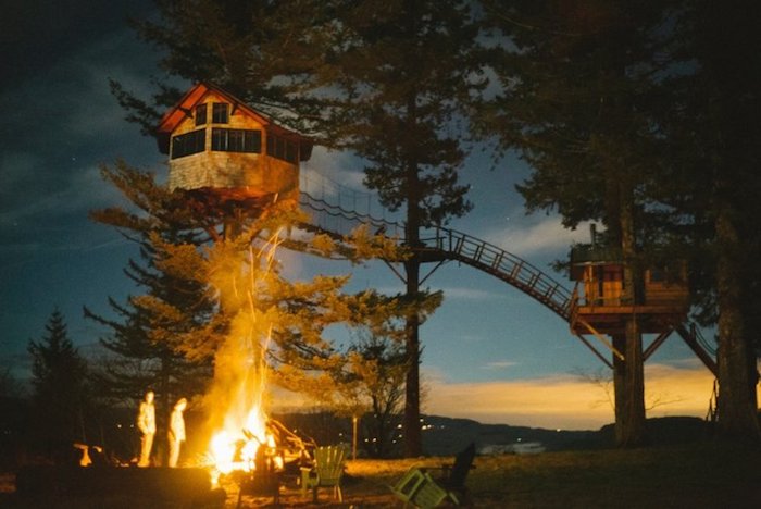 bonfire near two people, two tree houses built on large fir trees, and linked through a long bridge, photo taken at dusk