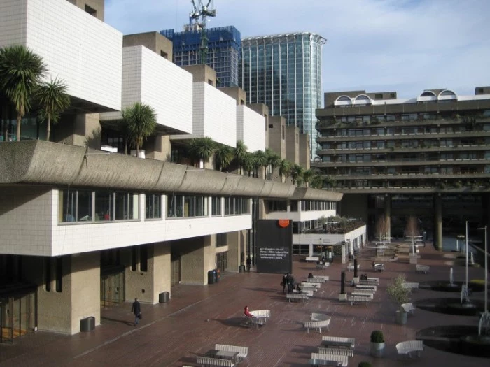 yard of the barbican centre, in london england, multiple white benches, near a building made of grey concrete, and decorated with white tiles