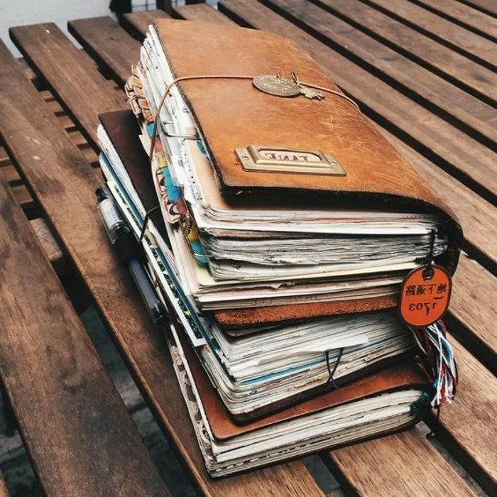 adventure journal, three big journals in light brown, with many colorful pages, stacked on top of each other, each tied with string, on a bench