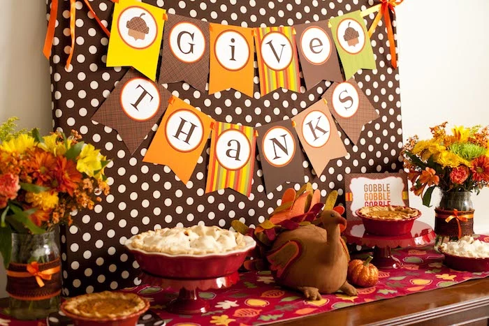 brown board with white polka dots, with a banner in yellow, orange, brown and green, spelling give thanks, near a wooden table with an autumn-leaf-patterned tablecloth, containing four pies in red dishes, a stuffed toy turkey, two decorated jars with yellow, orange and red flowers and a small pumpkin