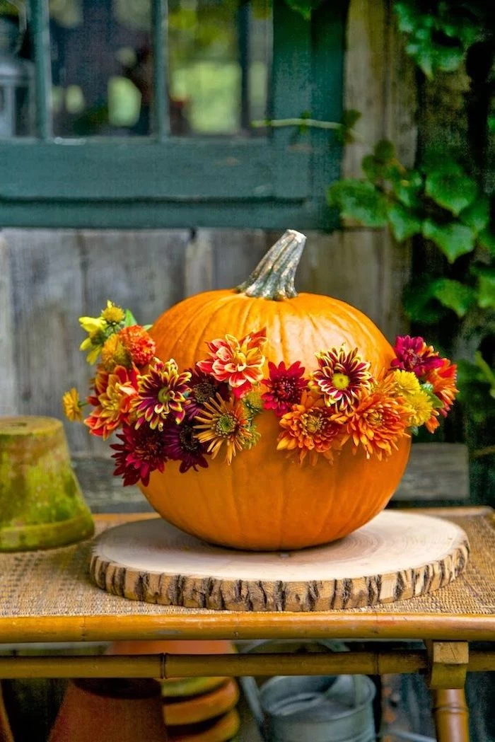 orange pumpkin, adorned with red, yellow, orange and green flowers, placed on a wooden mat, on a garden table, near planting pots, green window, plant and planks in the background