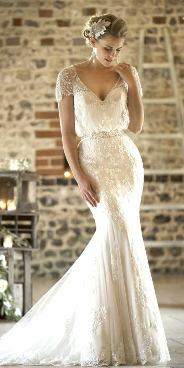 vintage inspired wedding dresses,young blonde bride looking down, wearing embroidered long white dress with lace sleeves and a hair ornament, brick wall and furniture in the background