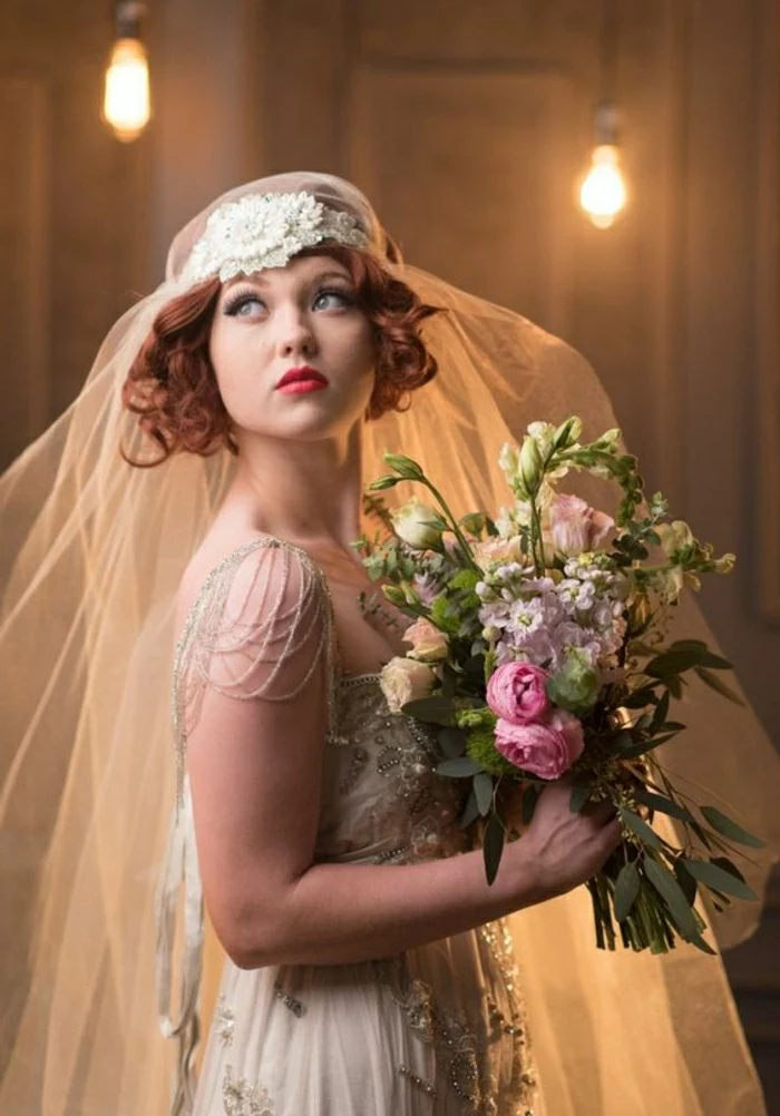 vintage inspired wedding dresses, woman with red lipstick and hair looking up, wearing vintage 1920s headdress and fail and bridal gown with lace beads and pearls, holding a large bouquet of differently colored flowers