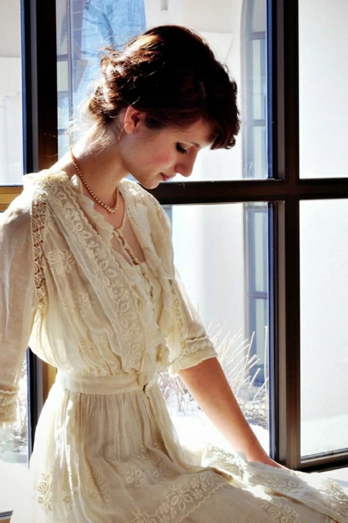 unique wedding dresses, woman with tied back dark hair, sitting near a window and looking down in profile, wearing a pearl necklace and a vintage cream Victorian era wedding dress with lace and embroidery