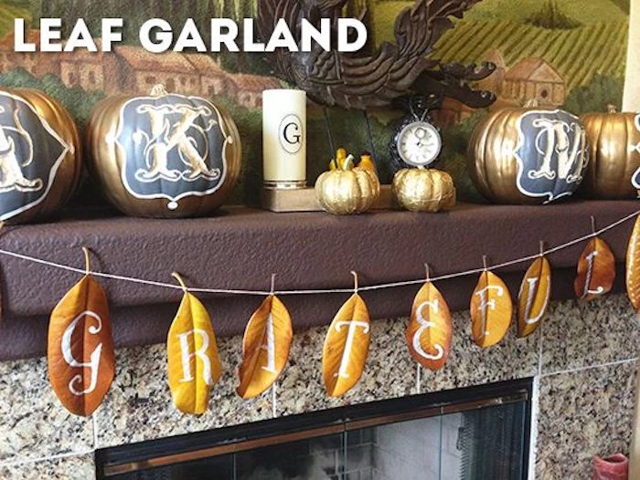 a garland made of yellow and orange autumn leaves spelling out grateful with white fancy letters,tied to a brown mantle containing four carved gold and blue pumpkins, two small golden pumpkins, a watch, a candle and a large painting, over a fireplace