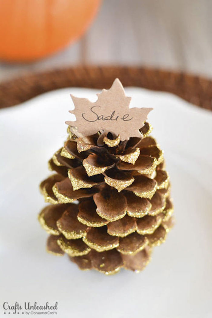 cute happy thanksgiving images, a pine-cone, painted with glittering gold paint, adorned with a paper leaf name card, placed on a white plate, sitting on a wooden mat on a table,