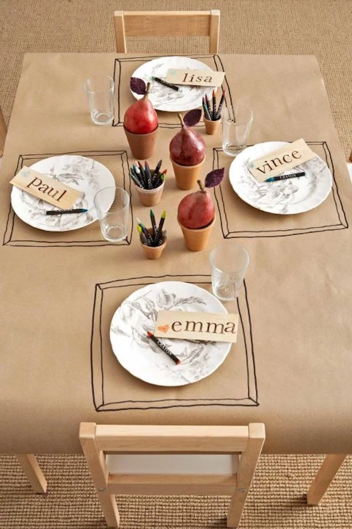 cute happy thanksgiving images, a paper tablecloth in sepia with table mats drawn by kids, four plates with name tags, three cups with crayons, four glasses, three cups holding red pears, two chairs