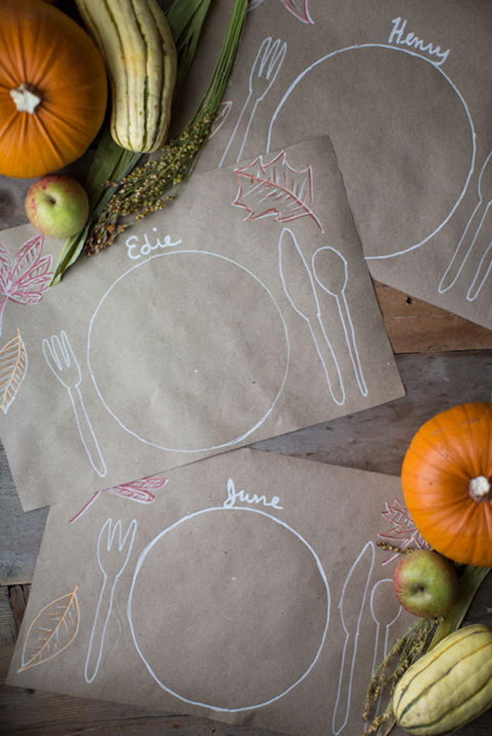 cute happy thanksgiving images, sepia paper mats, with drawings and names written by kids, on a wooden table, near pumpkins, gourds, apples and green plants