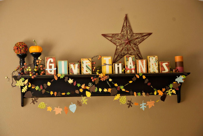 differently sized colorful wooden blocks spelling out give thanks, placed on a dark wooden shelf, adorned with a colorful candle, a round ornament, a small pumpkin and bright colorful garlands made of small paper leaves, on a light orange wall with a brown metal star
