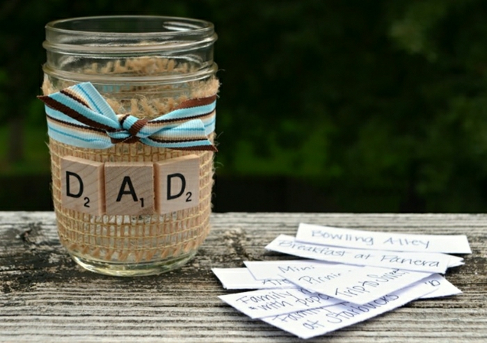 father's day homemade gifts, decorated jar with blue and brown ribbon, pieces of paper, placed on a wooden table, dark background