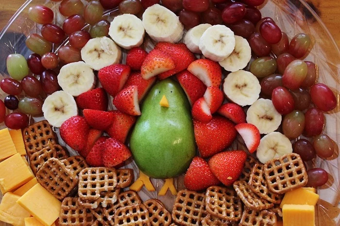 funny happy thanksgiving images, a pear, strawberry and banana slices, grapes, cheese slices and salted biscuits made to look like a turkey, placed on a clear plate, sitting on a wooden table