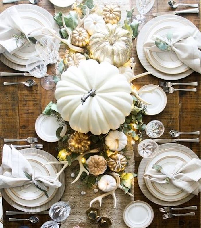 happy thanksgiving pictures, wooden table, white plates on white wooden mats, white napkins, silver cutlery, crystal wine and water glasses, burlap tablecloth, large white pumpkin, smaller pumpkins in gold and white, white antlers, green leaves decoration