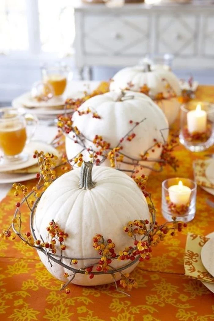 three white pumpkins, with wreaths made of twigs with orange berries, on a table with an orange and yellow tablecloth with white plates, glasses filled with orange juice and two small lit candles in clear glasses, white cabinets and window in the bakground 