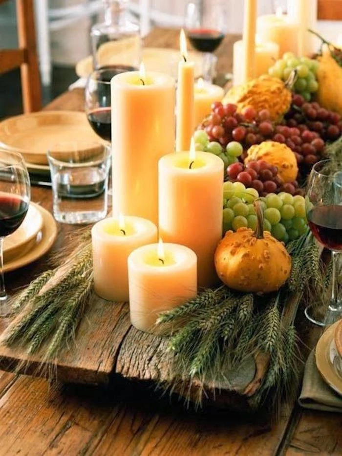 happy thanksgiving pictures, lit candles in different shapes and sizes, placed on a rough wooden aboard, next to grapes, green wheat bunches, gourds, little pumpkins, on a wooden table with plates, full wine and water glasses