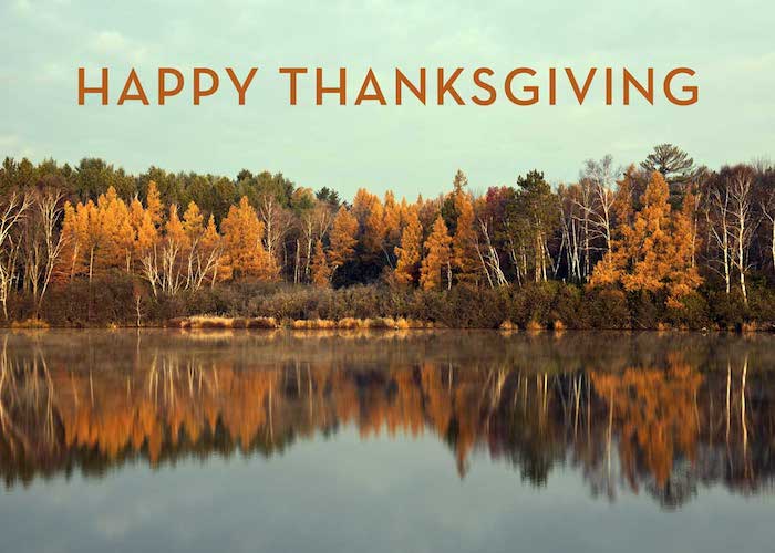 images of thanksgiving, forest in autumn with orange and green trees, blue sky with clouds, all reflected in a lake, brown writing saying happy thanksgiving