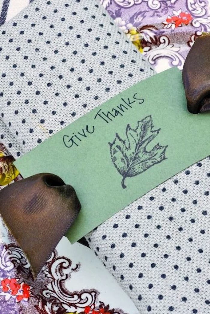 napkin made of light blue fabric with dark blue polka dots, with a brown ribbon and a green label with a stamp and the writing give thanks, on a colorful tablecloth