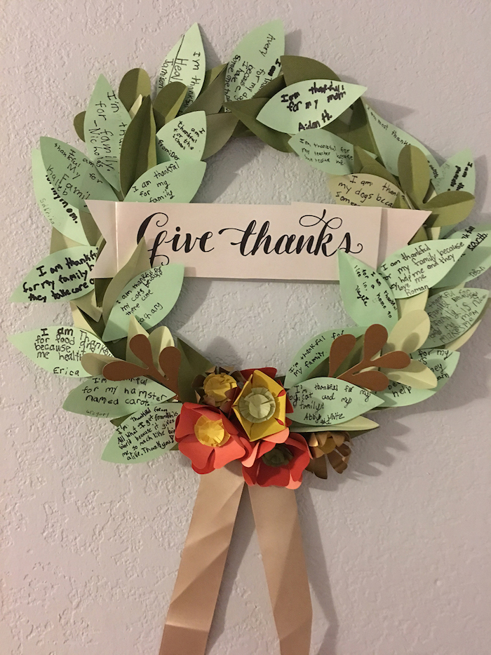 wreath made of green paper leaves with messages, with white banner saying give thanks, orange, red and yellow paper flowers and light brown ribbon, light grey background