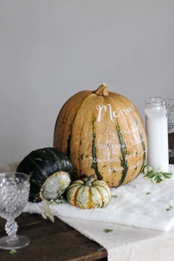 large orange and green pumpkin with a dinner menu written in white, two smaller pumpkins in light and dark green, all placed on a white table cloth, on a wooden surface with two crystal glasses and a large white candle