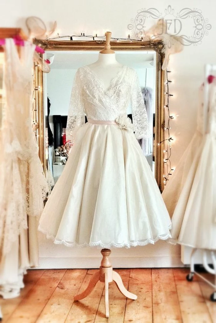 short lace wedding dress, dress with lace top and pink belt with flower, sheer sleeves and plain bottom, on a simple wooden mannequin, in front of a mirror with fairy lights in a room with other white dresses 