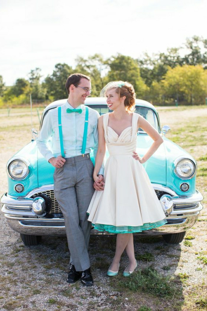 short white wedding dresses, a man with glasses in white shirt and turquoise bow tie and overalls, and a woman in calf-length dress with turquoise petticoat and shoes, holding hands in front of a turquoise retro car