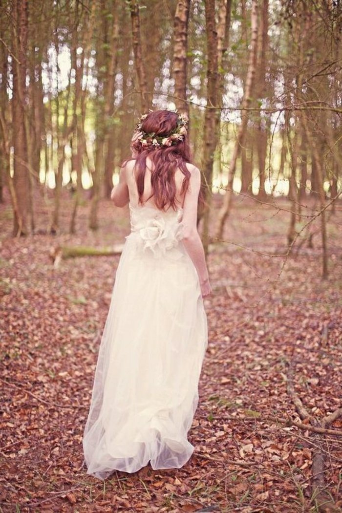 unique wedding dresses, auburn-haired bride in long dress made of tulle, facing backwards and walking in the woods, one hand holding a rose crown on her head, trees, leaves
