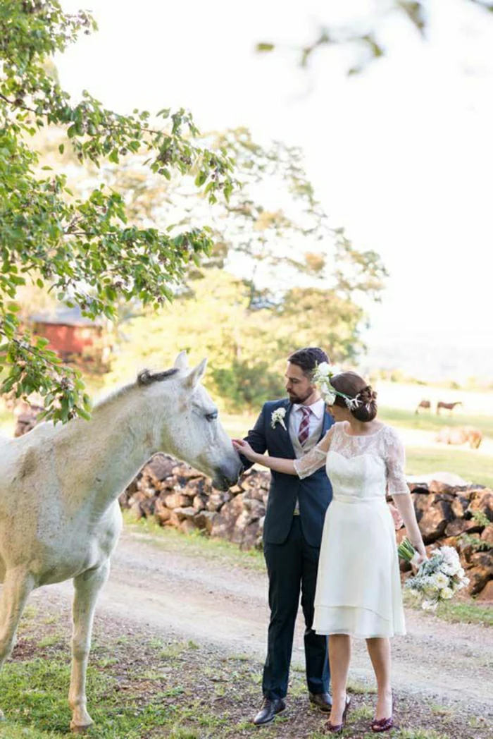 short white wedding dresses, bride in white knee-length dress with lace details and a head ornament with white flowers, holding a bouquet and a groom in black suit and red-striped tie petting a white horse near a field and trees