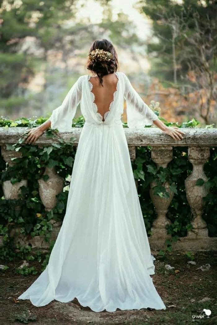 simple wedding dress, woman in long white dress with wide sleeves and an open back, leaning on a terrace made of stone with green creeping plants and trees in background, messy hair bun with ornaments