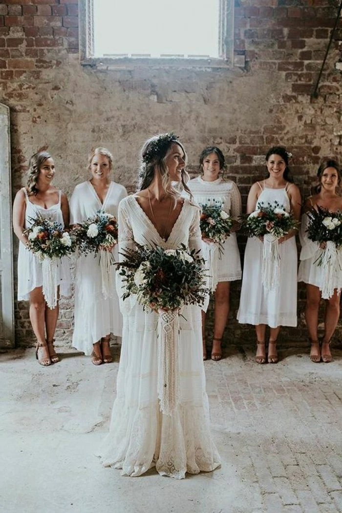 simple wedding dress, smiling bride in white long bohemian hippie dress holding a large green and white bouquet, five bridesmaids in white dresses holding flowers in background, old brick shabby interior