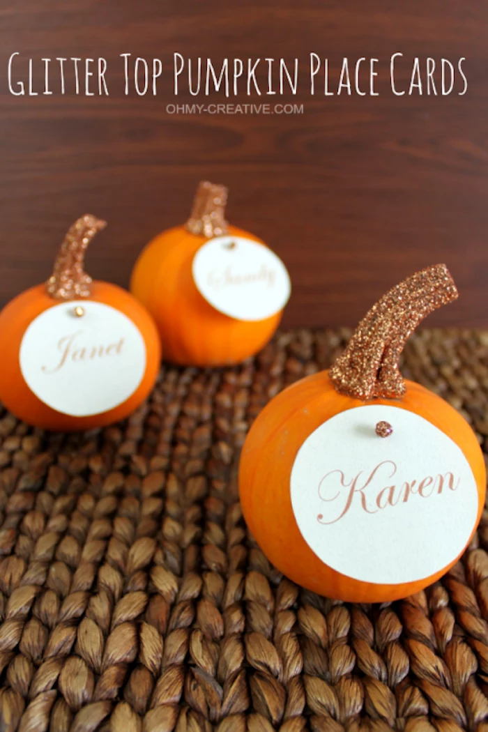 three small orange pumpkins, stems painted with glittering golden paint, with round name tags attached with a pin, on a rough fabric surface, wooden background with white writing