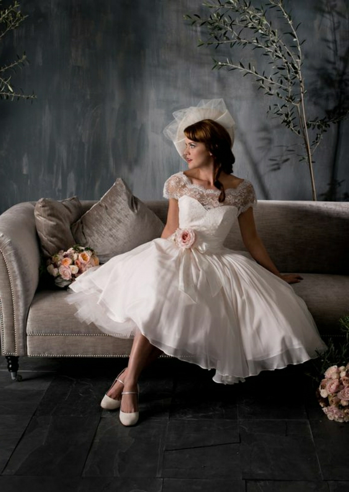 tea length wedding dresses, brunette woman in bridal dress and hat sitting on a grey couch, lace details and a pink rose on her belt, two bouquets of roses, grey background with small decorative tree