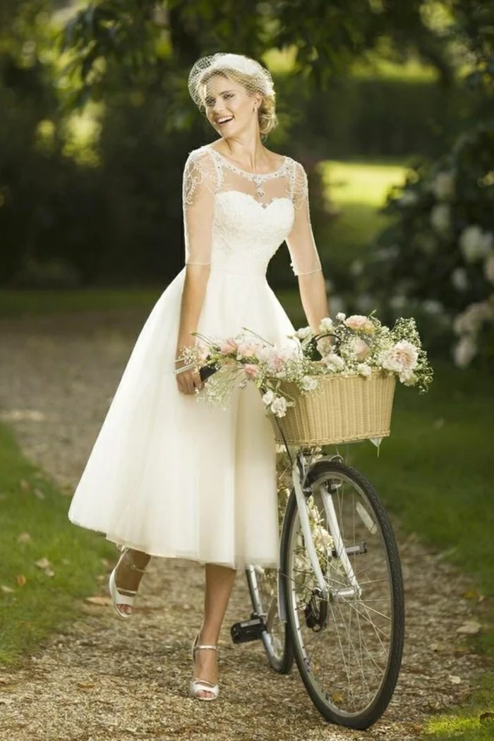 tea length wedding dresses, smiling bride in calf-length white dress with sheer sleeves pushing a bike with a flower basket 