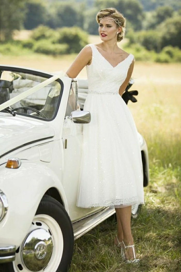 tea length dresses, fair-haired woman in simple wedding dress, with vintage-inspired hairstyle and a forehead tiara, leaning on a white retro car with bow, green fields in background