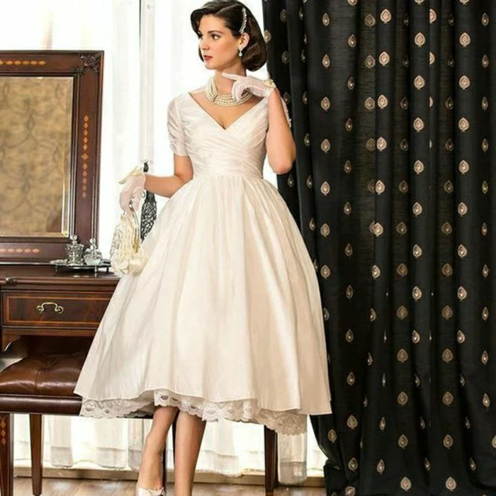 tea length dresses, bride in simple 1950s calf-length wedding dress, with white short gloves pearl necklace airings and hair ornament, holding a white purse,, vintage furniture and a dark curtain