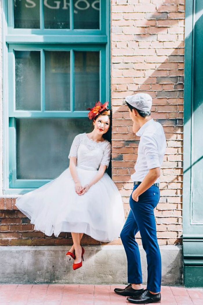 short white wedding dresses, young bride with simple white dress red shoes and red hair ornament, sitting on a window sill and looking at a young man in jeans, white shirt and grey cap