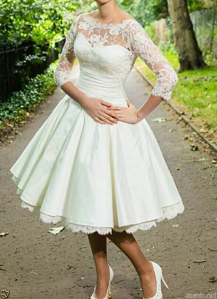 tea length dresses, close up of a woman's body in white calf-length bridal dress with lace details and sheer sleeves, hands on stomach and hips and wearing white shoes, park in background 