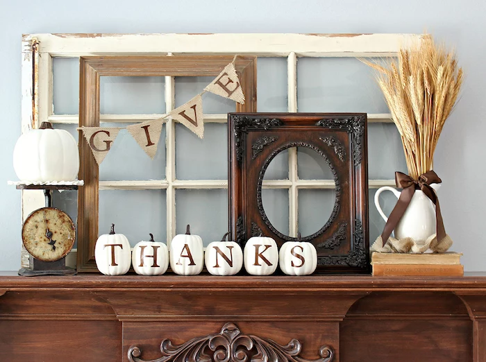 thanksgiving photos, a wooden carved mantle, with five little white pumpkins spelling out thanks in brown letters, a milk jug placed on two books, with a bunch of wheat tied with a brown ribbon in it, three wooden frames in different colors with a banner saying give, a bigger white pumpkin and decorations