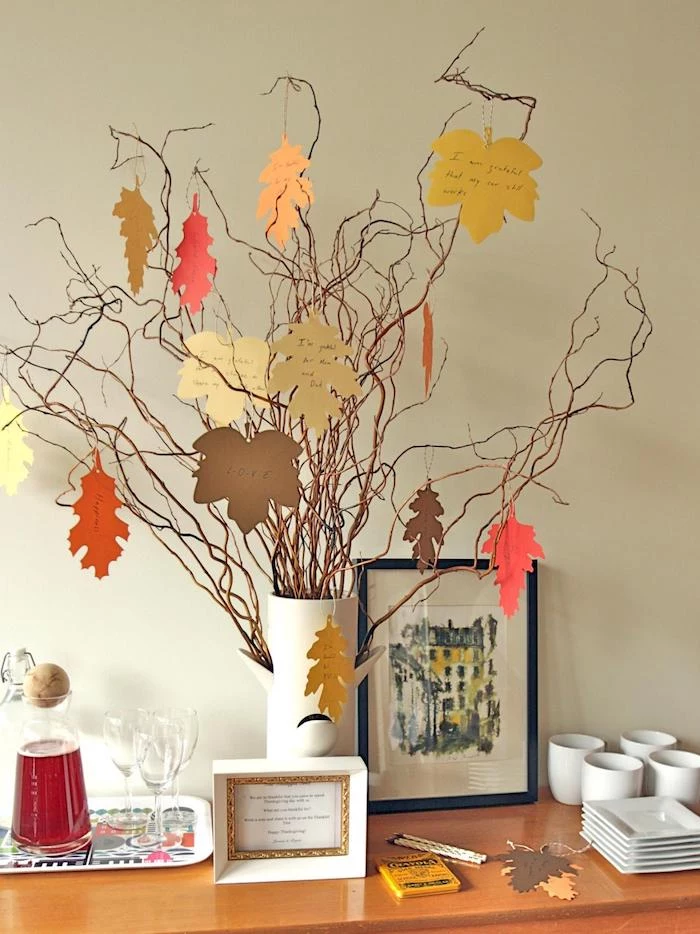 thanksgiving photos, dried tree branches, adorned with yellow, orange and brown leaf-shaped cutouts with messages, in a white vase, on a wooden table, with pitcher of juice and glasses on a tray, white coffee cups and square plates and two frames