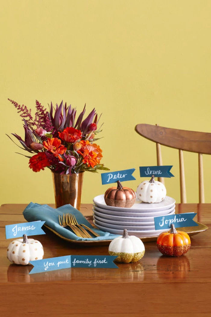 thanksgiving photos, little painted pumpkins in various colors, with blue name tags attached to their stems, on a brown wooden kitchen table, featuring several white plates and a blue napkin with silver forks, on a tray, near a golden vase with red autumn flowers, chair, yellow background