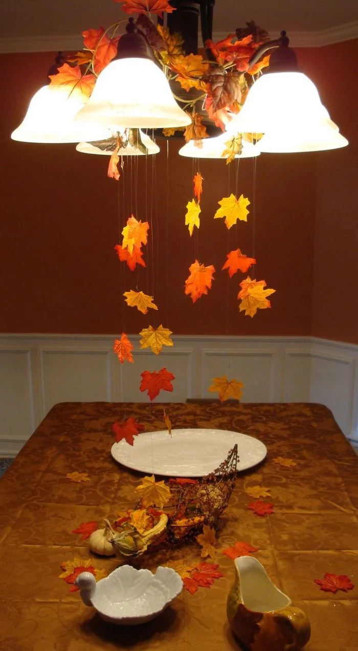thanksgiving pics, yellow and orange leaves, hanging on strings, from a lit chandelier, over a brown kitchen table, with s white plate, gravy jug, swan shaped bowl, and a cornucopia wire ornament with small pumpkins, gourds and autumn leaves, white and dark background
