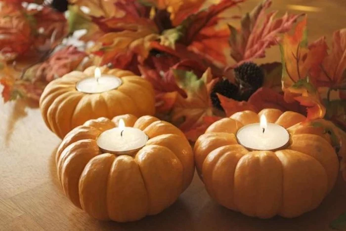 thanksgiving pics, three orange pumpkins wit lit white candles inside, on a wooden surface, colorful autumn leaves in the background 