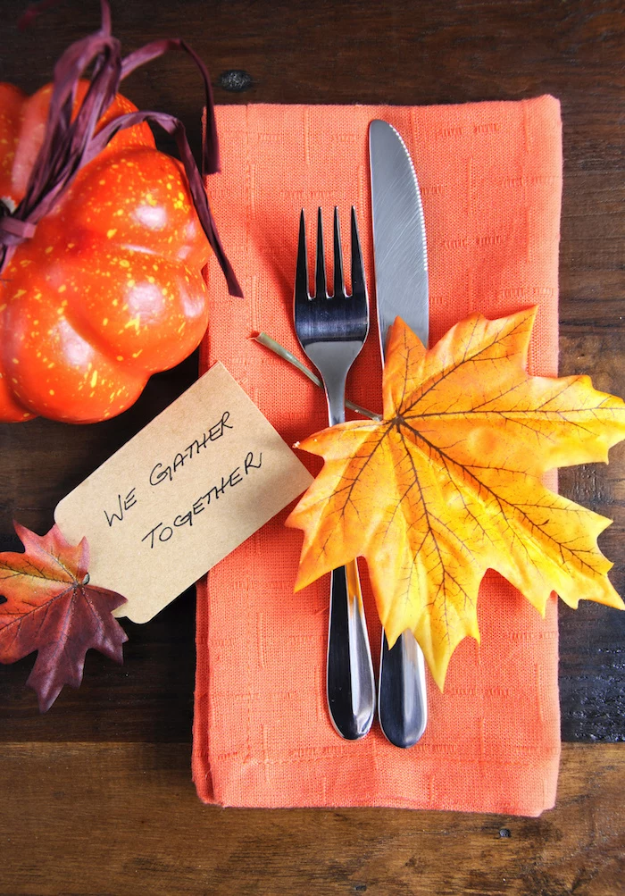 thanksgiving pics, wooden table with an orange pumpkin decoration tied with violet straw cord, next to an orange napkin with silver fork and knife, decorated with a big yellow leaf and a small colorful leaf attached to a piece of paper with a festive message
