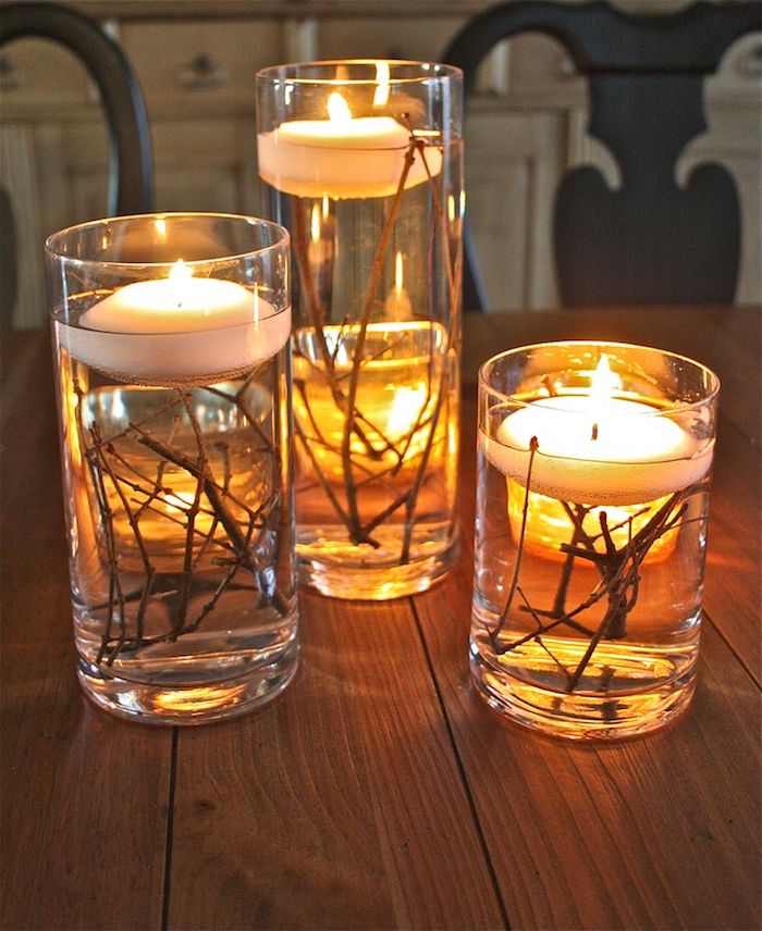 glasses of different sizes filled with water and twigs with lit floating candles inside, on a wooden table with two chairs and white cabinets in the background