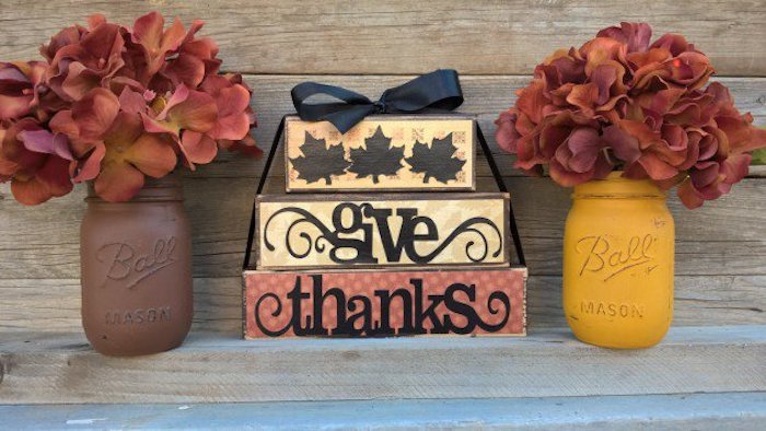 one orange and two yellow boxes, stacked on top of each other, tied with a black ribbon, featuring leaves and the words give thanks, placed on wooden steps, near two mason jars painted in brown and yellow and containing red flowers