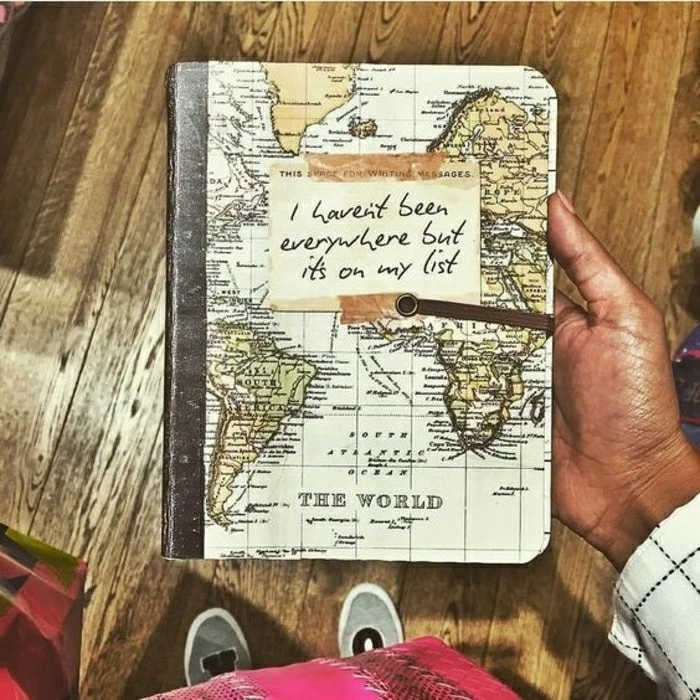 travel journal, a tan hand holding a notebook, with a yellow and green map of the world on the cover, shoes, wooden boards