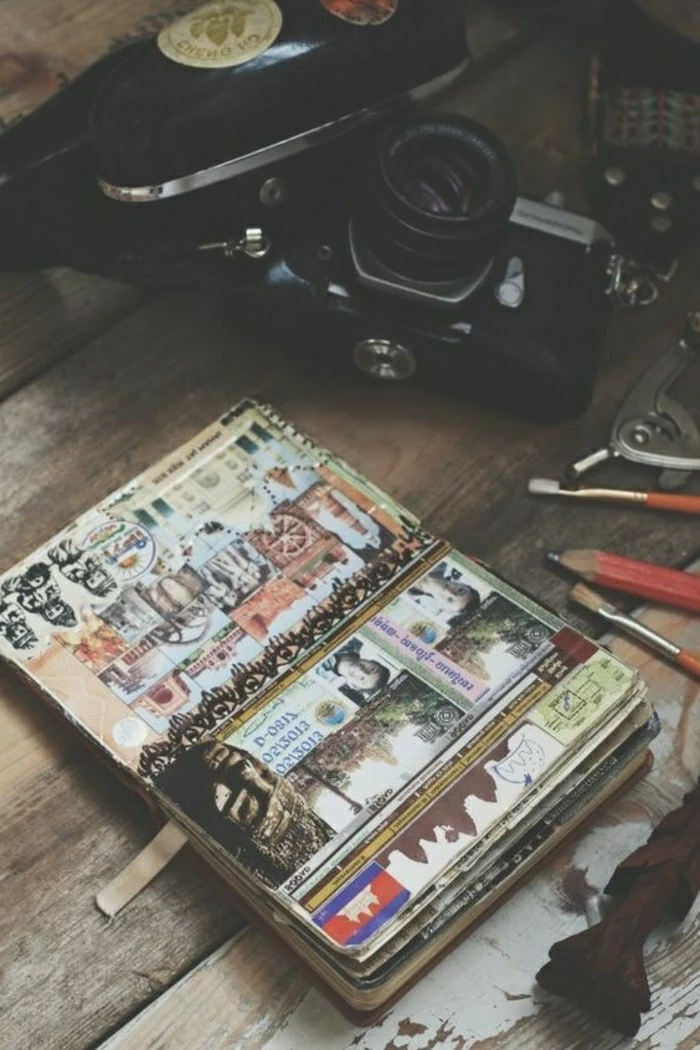 travelogue, wooden surface, open notebook with various photos, cutouts, stickers, drawings, collage,black camera, pencils, paint brushes