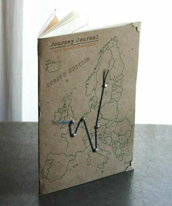 travelogue, light brown notepad with map outlines and blue thread pinned on cover, recycled paper, on dark surface and light background