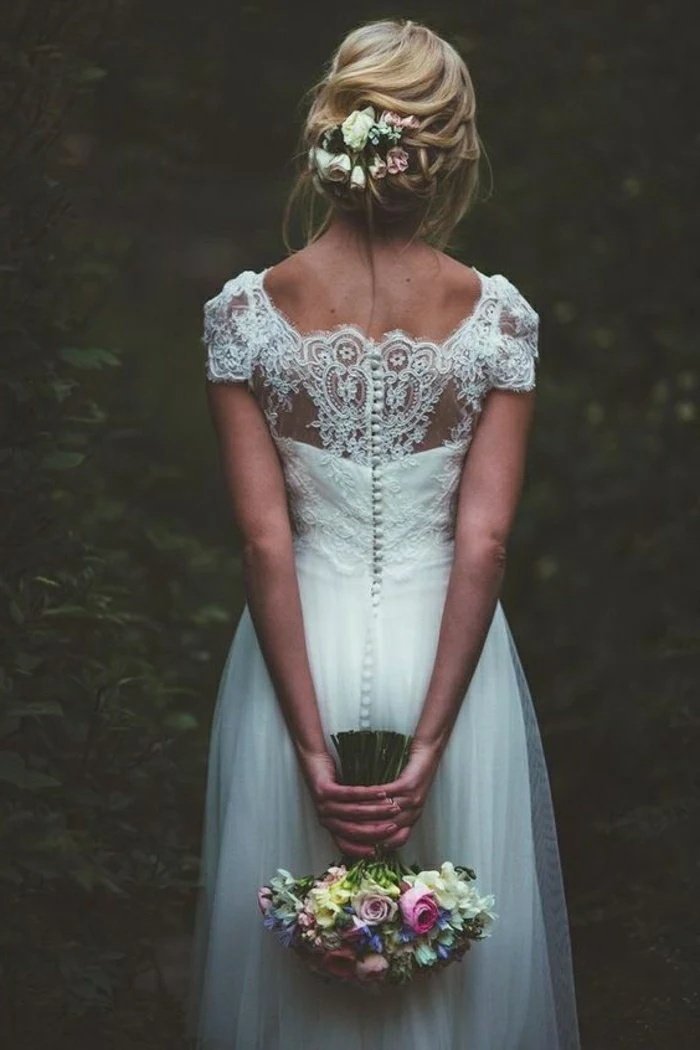 vintage wedding dresses, blonde bride with flowers in hair facing backwards, wearing a long white bridal dress with lace back and buttons, holding a bouquet of flowers, dark forest background