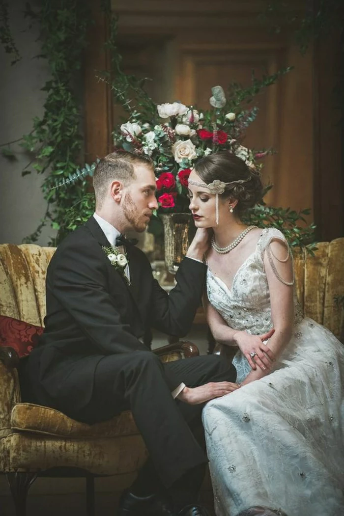 vintage inspired wedding dresses, bride and groom sitting and hugging on a yellow couch, woman wears a 1920s style dress with embroidery beads and pearls and a hair ornament, man wears black suit, flowers in background